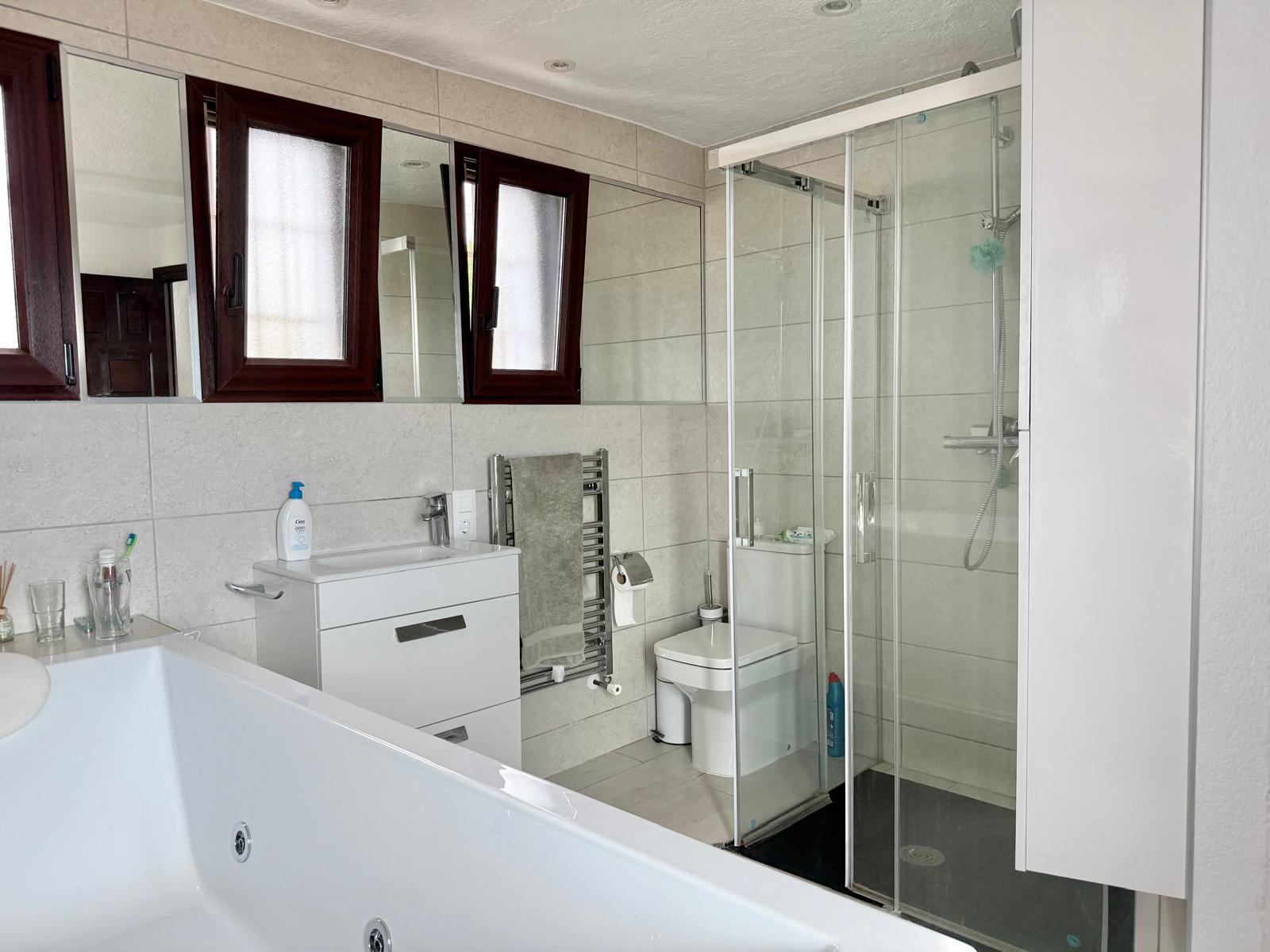 Baño equipado con bañera de hidromasaje, ducha independiente con mampara de cristal, mueble lavabo blanco y radiador toallero bajo una iluminación empotrada.