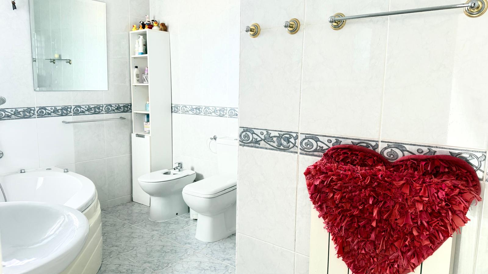 Baño equipado con bañera de hidromasaje en esquina, bidet e inodoro, con paredes alicatadas en blanco y una cenefa decorativa clásica.