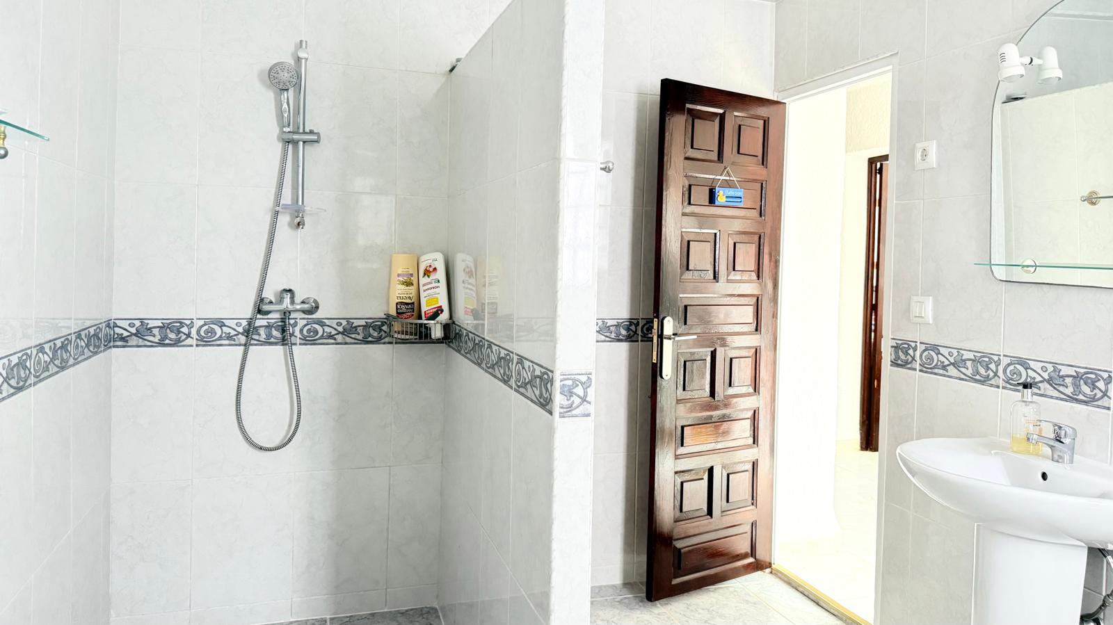 Baño funcional con zona de ducha alicatada, cenefa decorativa clásica y puerta de madera maciza tallada. Incluye lavabo de pedestal y espejo con iluminación superior.