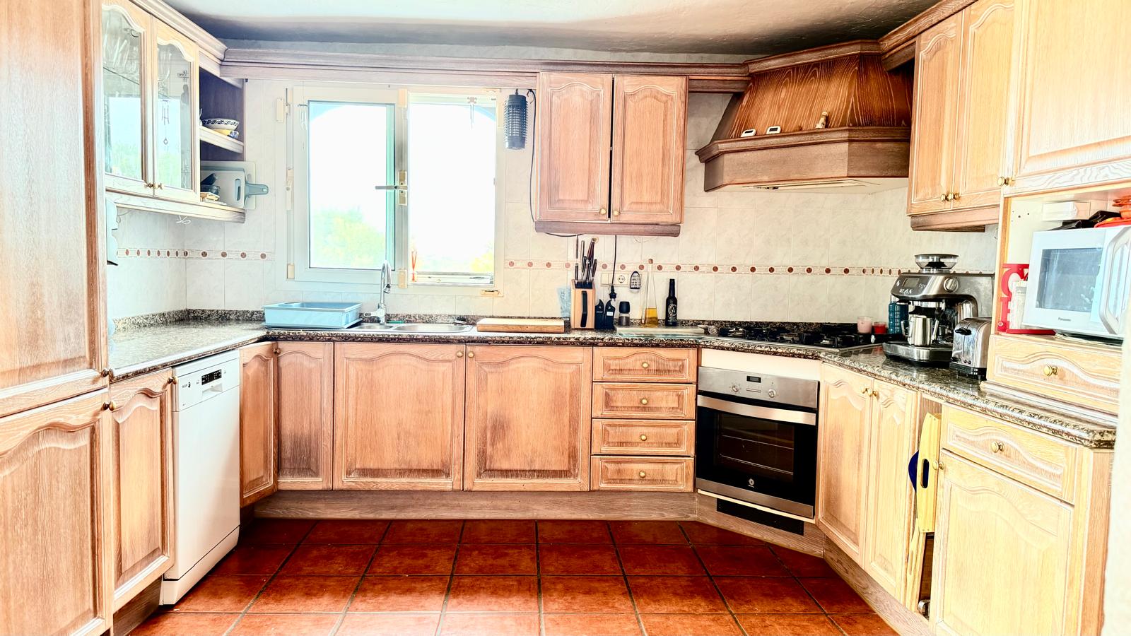 Cocina de estilo tradicional con mobiliario de roble, encimeras de granito y suelos de terracota. Incluye electrodomésticos integrados y una amplia campana extractora de madera.