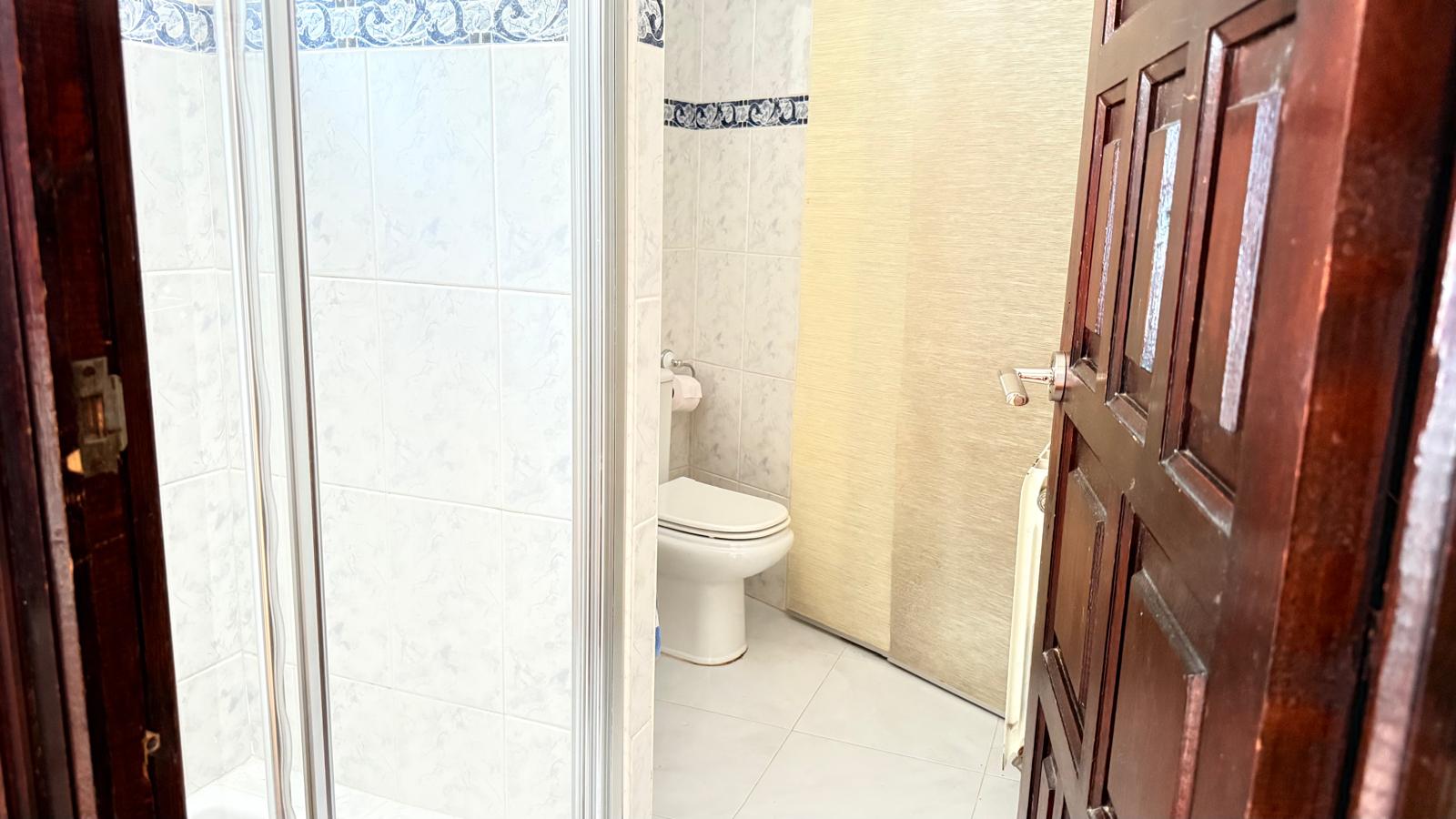 Cuarto de baño con azulejos blancos y cenefa decorativa azul, equipado con inodoro blanco y ducha con mampara de cristal, accesible mediante una puerta de madera maciza.