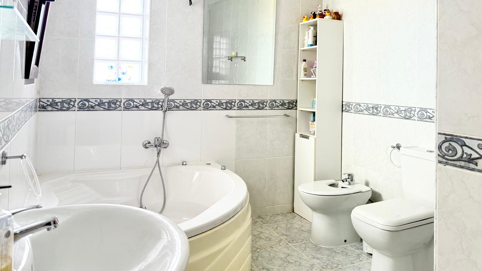Cuarto de baño con bañera de hidromasaje en esquina, bidet e inodoro. Presenta azulejos blancos con cenefa decorativa y luz natural a través de una ventana de pavés.