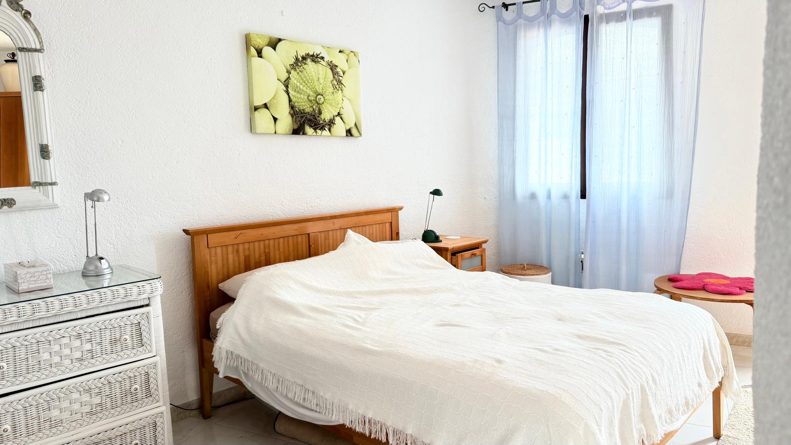 Dormitorio con cama de madera, cómoda de mimbre blanco y paredes de yeso texturizado. La estancia cuenta con luz natural filtrada por cortinas azules y suelos cerámicos.