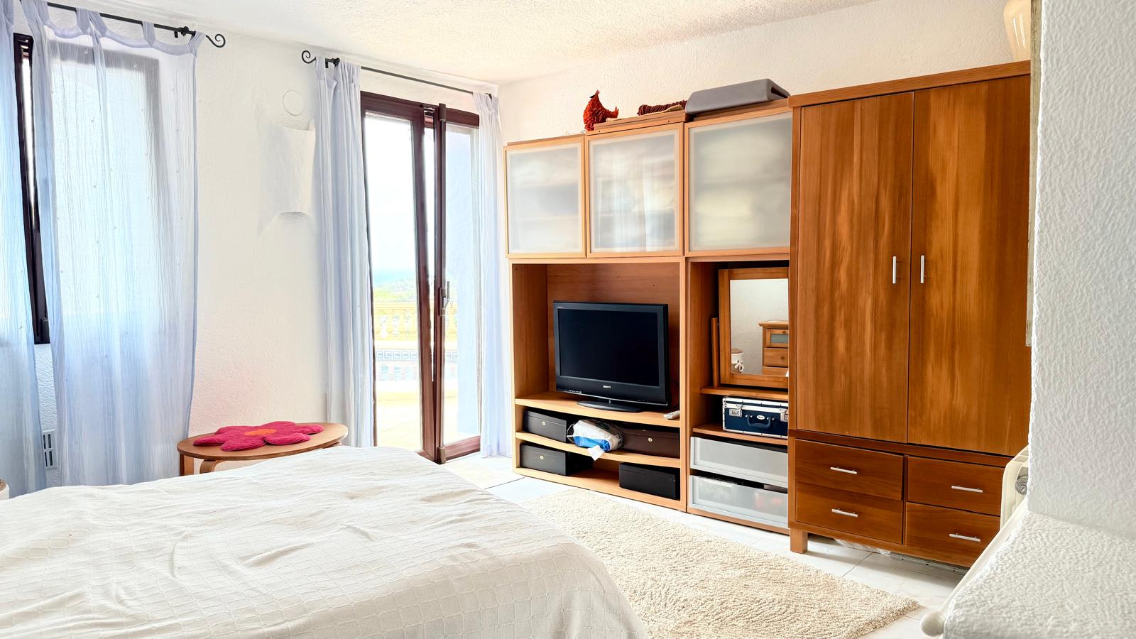 Dormitorio luminoso con acceso directo a la terraza, mobiliario de madera integrado con zona de TV y vistas despejadas a través de los ventanales.