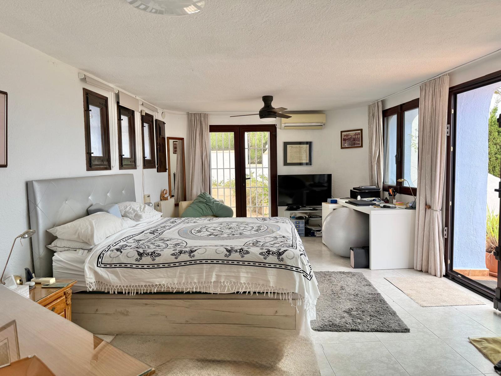 Dormitorio principal amplio con zona de despacho, aire acondicionado y ventilador de techo. Dispone de acceso directo a la terraza y ventanales con marcos de madera oscura.