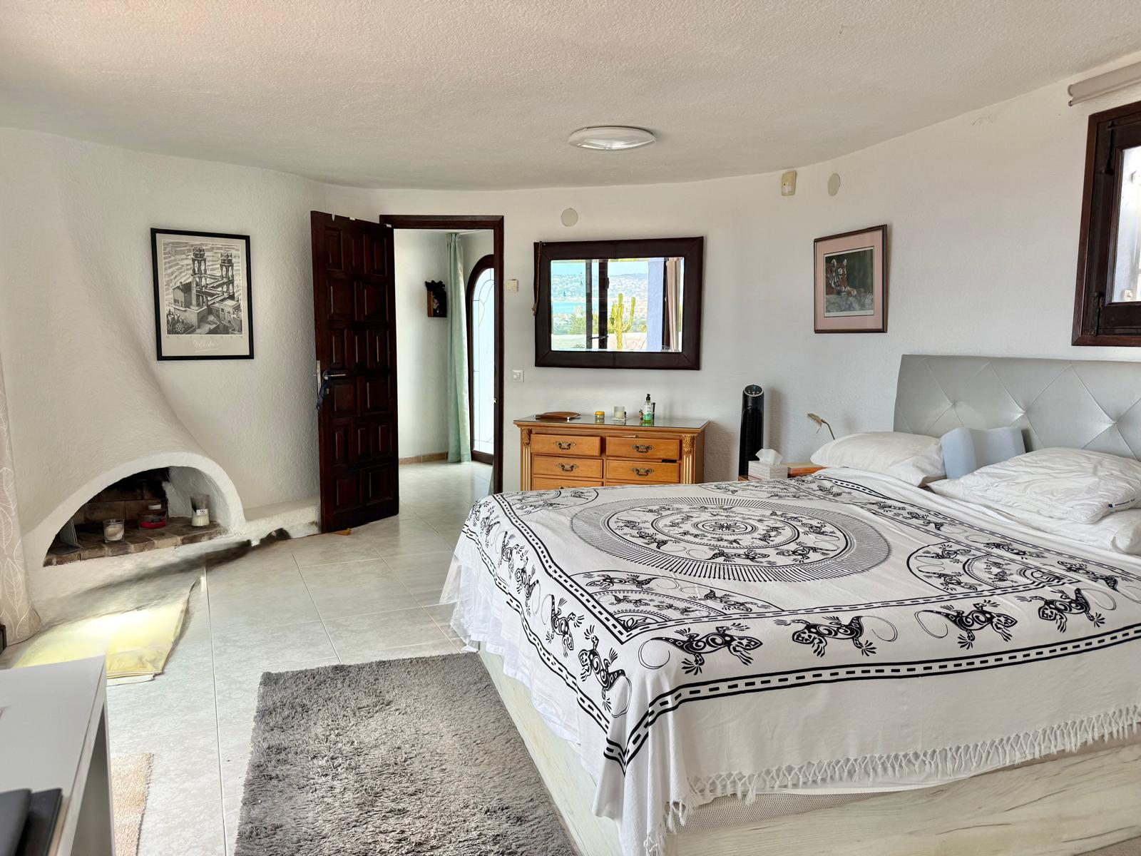 Dormitorio principal de estilo ibicenco con chimenea de obra curva, suelos cerámicos y un espejo que refleja las vistas panorámicas al mar de Jávea.