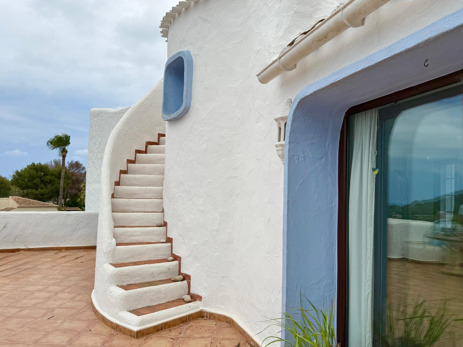 Escalera exterior curva de estilo ibicenco con peldaños de barro, integrada en la fachada blanca con detalles en azul y suelos de terracota.