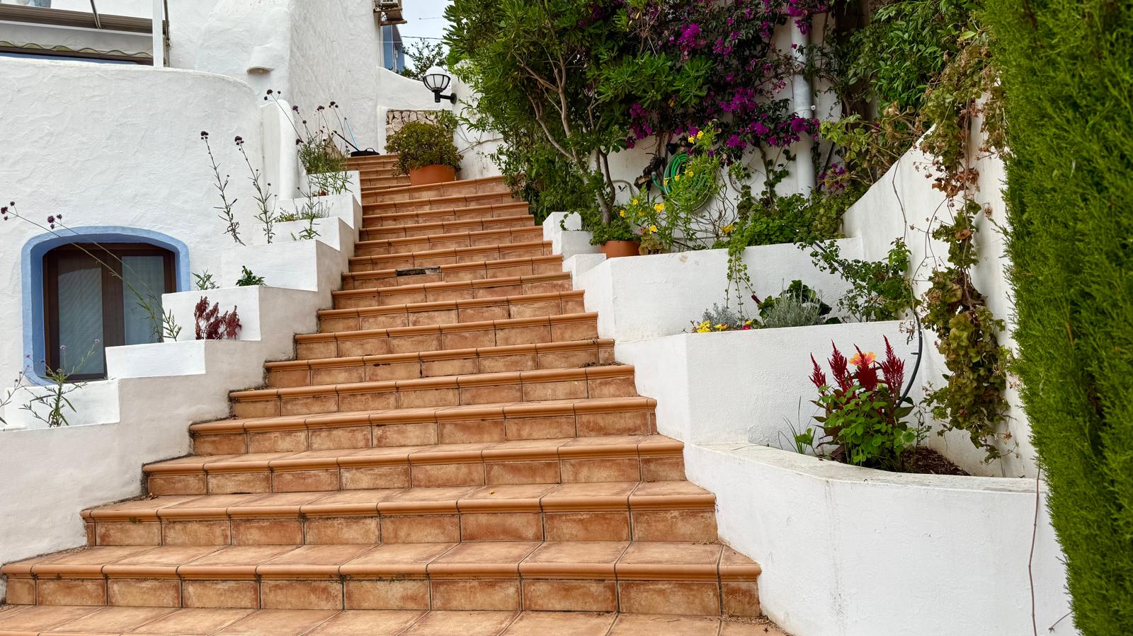 Escalera exterior de baldosas cerámicas que conecta los niveles de la villa, flanqueada por jardineras blancas integradas con vegetación mediterránea y flores.