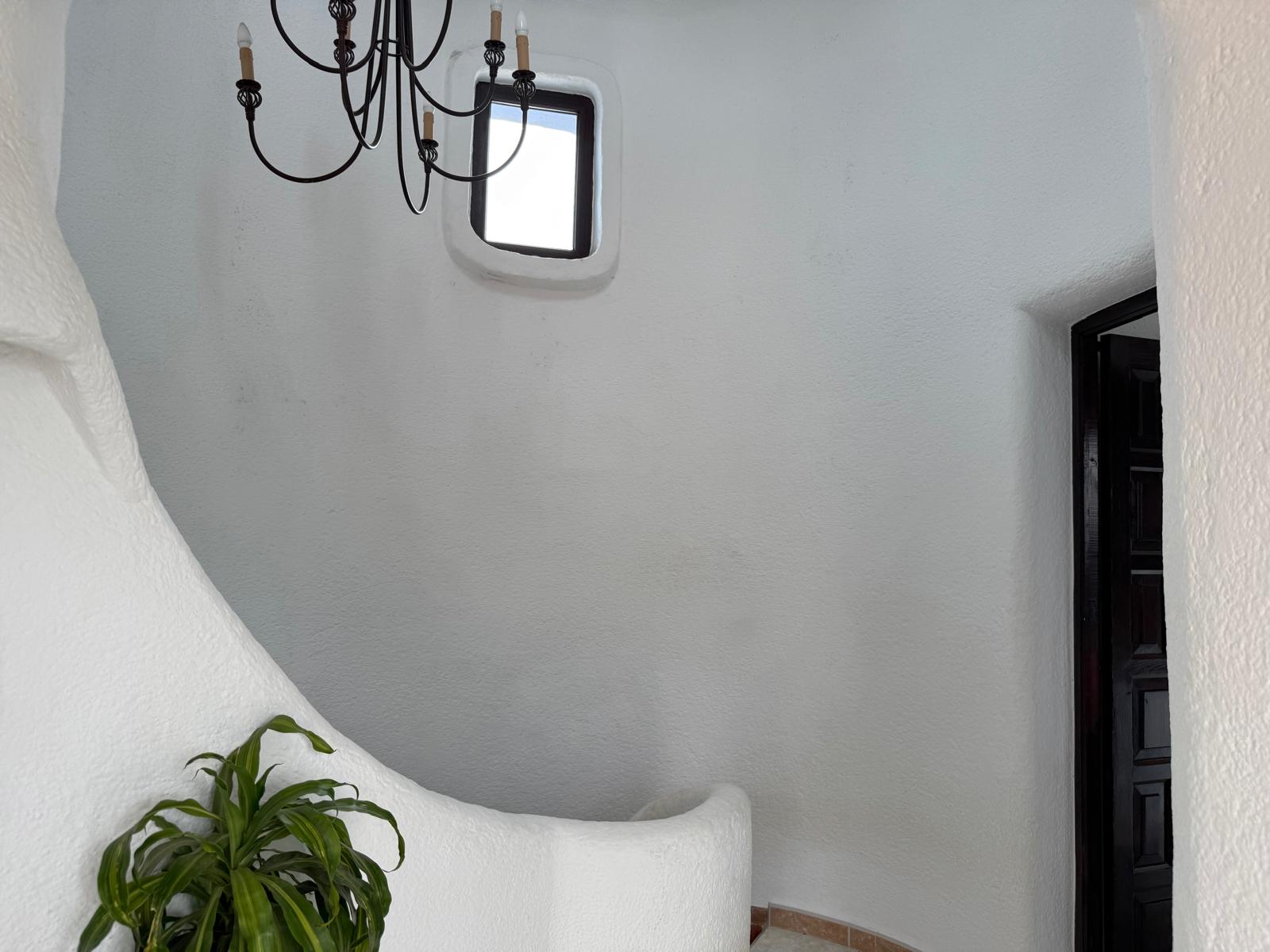 Escalera interior curva de estilo ibicenco con paredes de yeso blanco texturizado, lámpara de araña de hierro forjado y una pequeña ventana con luz natural.