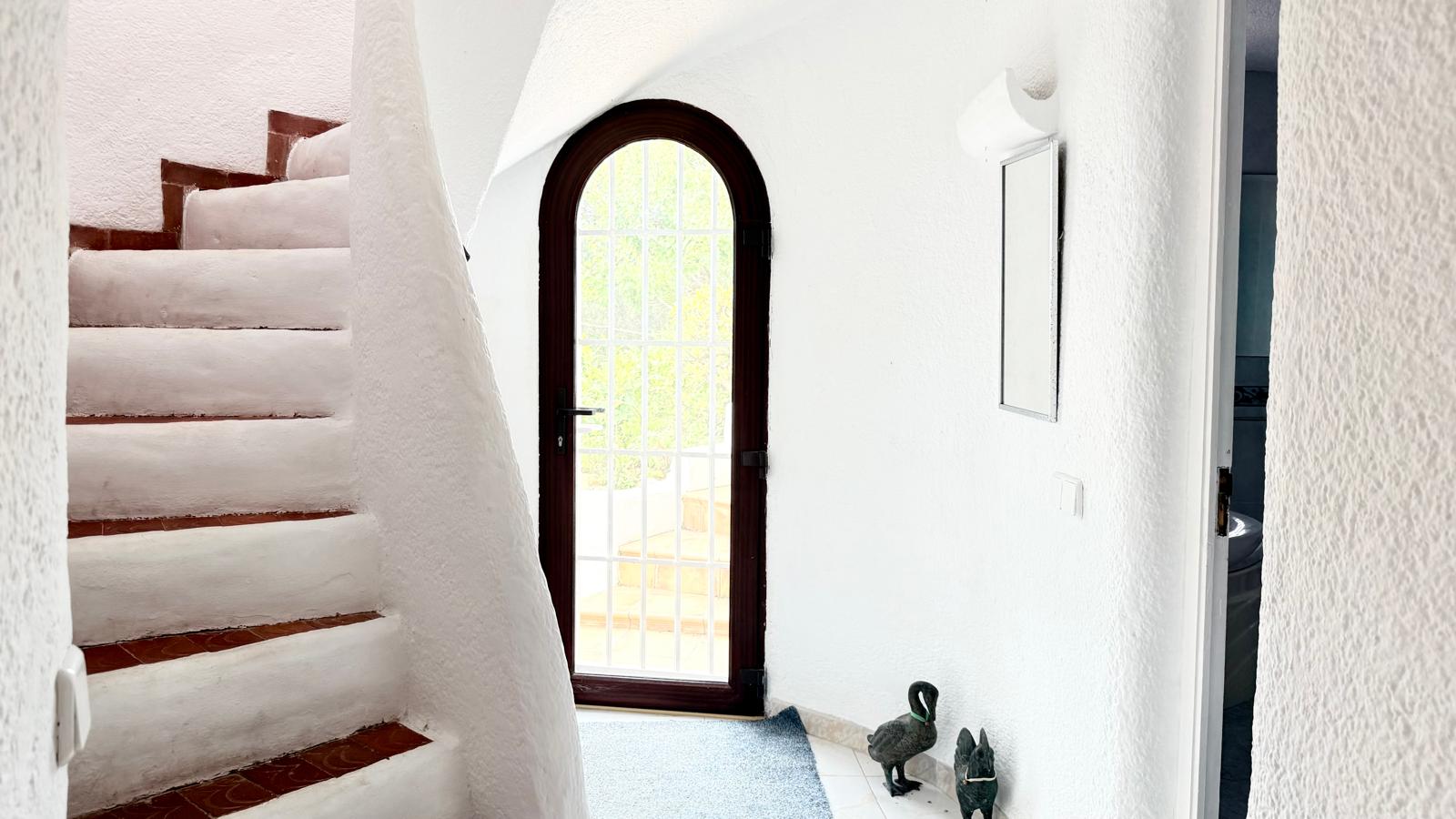 Escalera interior curva de estilo ibicenco con peldaños de barro cocido y paredes blancas texturizadas que conducen a una puerta de arco con acceso al exterior.