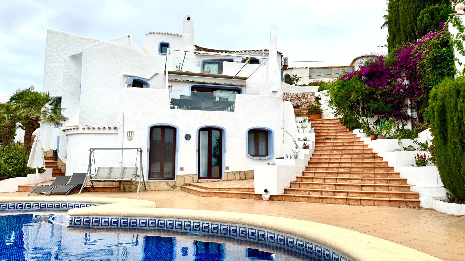 Fachada blanca de estilo ibicenco con carpintería azul, terraza de baldosas cerámicas y amplia escalera que conecta los niveles junto a la piscina y el jardín.