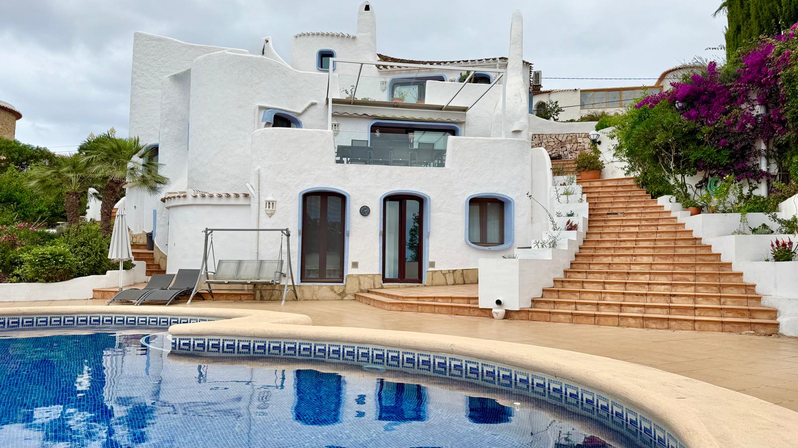 Fachada blanca de estilo ibicenco con formas orgánicas, amplias escaleras de terracota y zona de piscina rodeada de vegetación mediterránea.