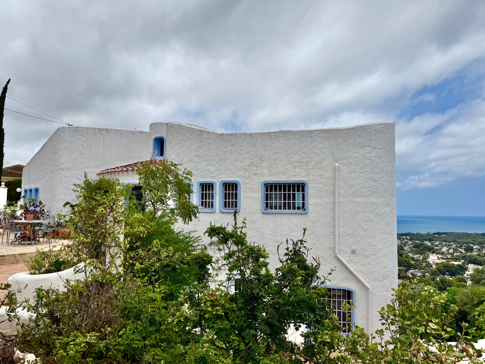 Fachada blanca de estilo ibicenco con ventanas de marcos azules y líneas curvas, situada frente a una vista panorámica del mar Mediterráneo.