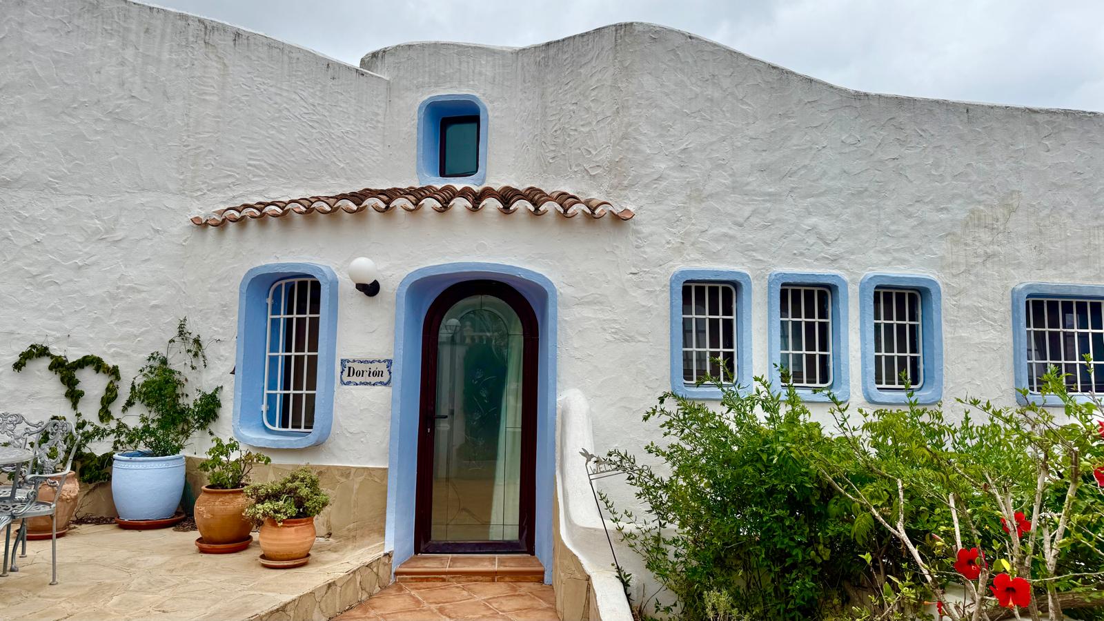 Fachada de estilo ibicenco con muros blancos, marcos de ventanas en azul y puerta de arco acristalada. Incluye suelos de barro y vegetación mediterránea en macetas.
