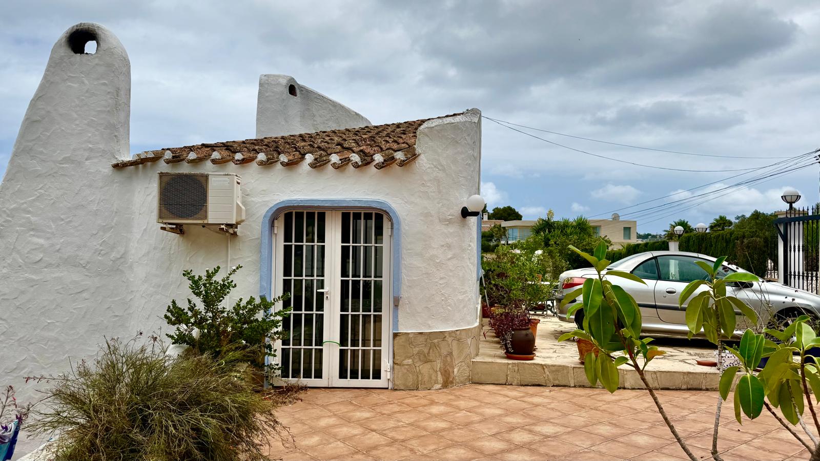 Fachada de estilo ibicenco con muros blancos redondeados, chimeneas tradicionales y zócalo de piedra. Incluye zona de aparcamiento nivelada y terraza con suelos cerámicos.