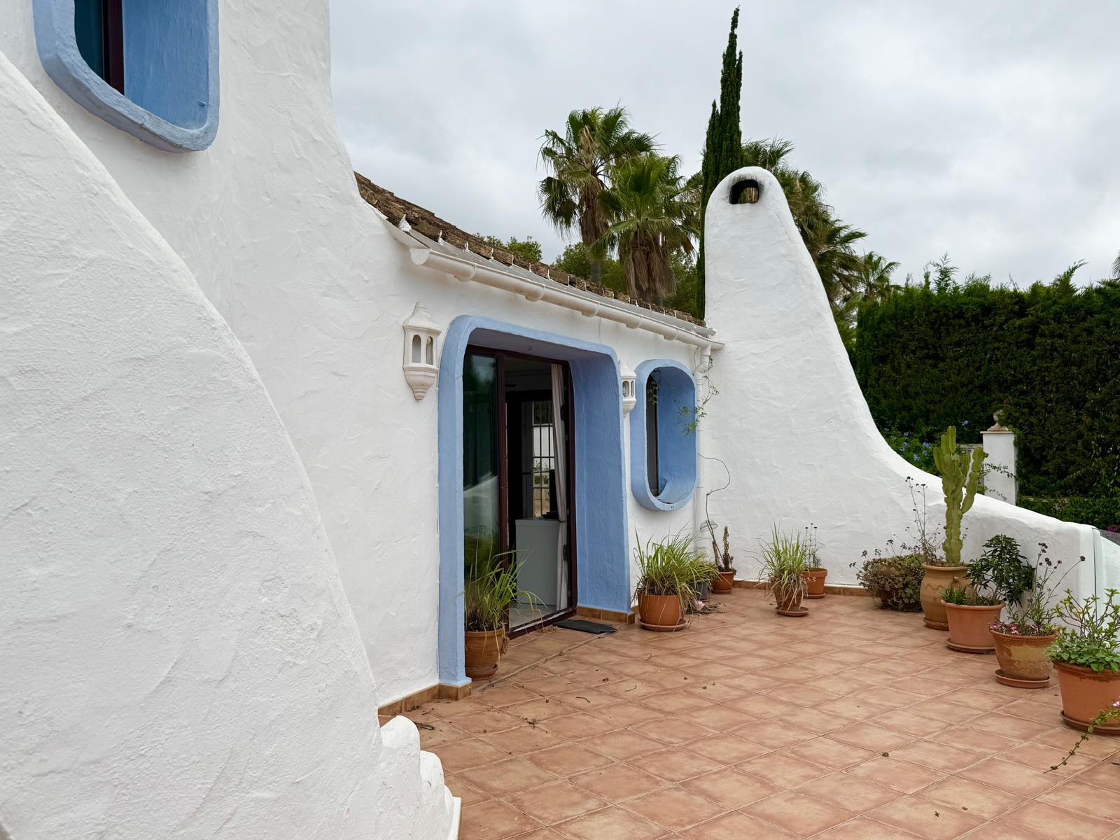 Fachada de estilo ibicenco con muros blancos redondeados, marcos azules y una terraza de baldosas de barro con plantas mediterráneas y una chimenea escultórica.