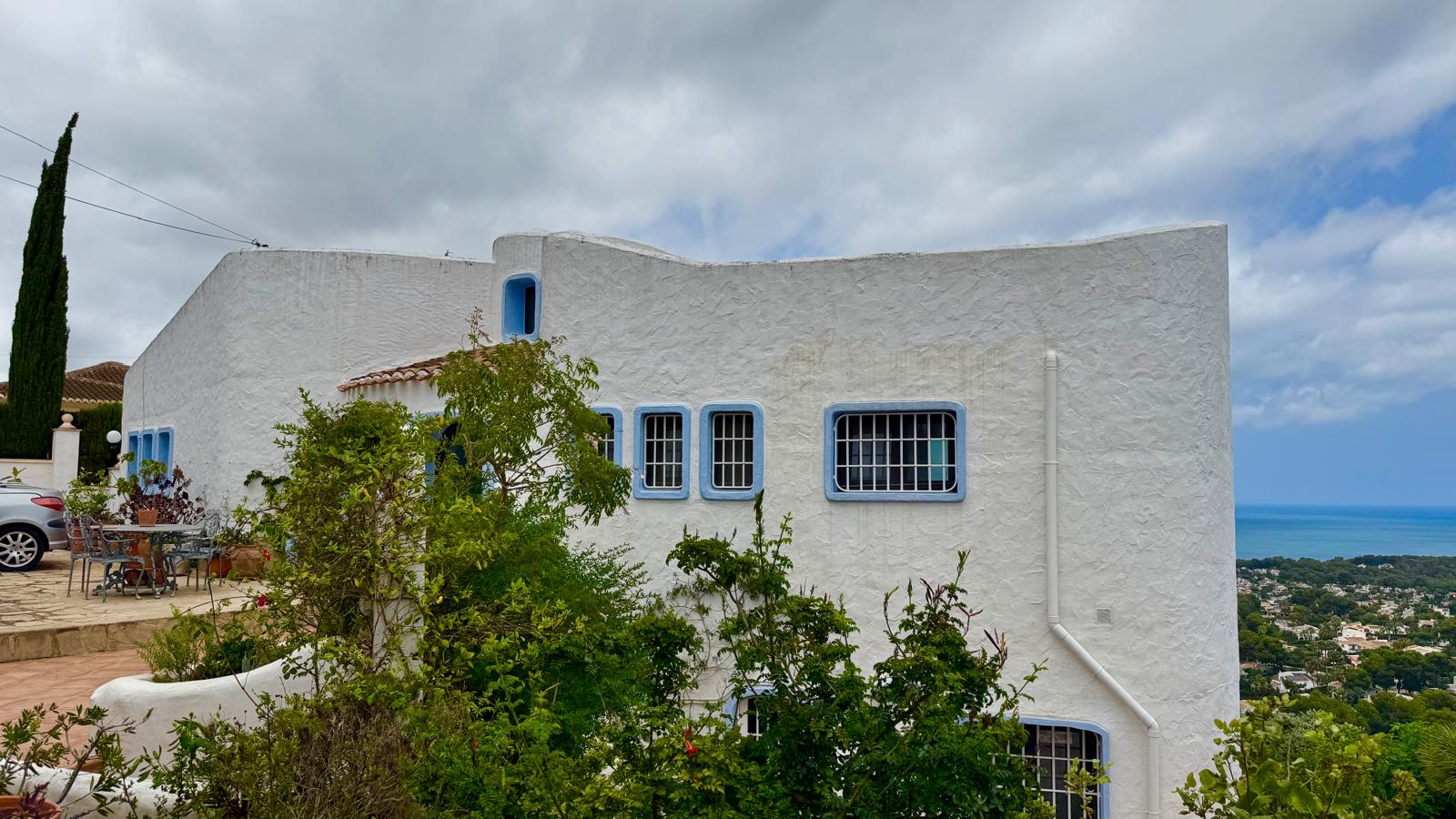 Fachada de estilo ibicenco con muros blancos texturizados, carpintería azul y líneas curvas orgánicas, situada en una posición elevada con vistas panorámicas al mar.