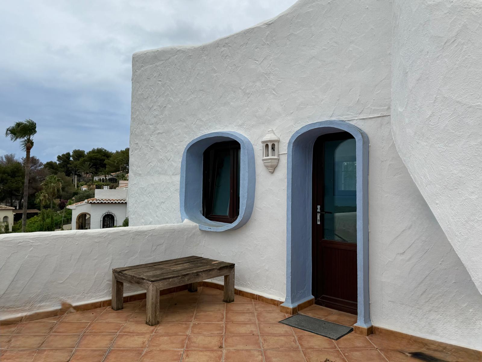 Fachada de estilo ibicenco con muros blancos texturizados, carpintería de madera oscura y marcos arqueados en azul. La terraza cuenta con suelo de terracota y una mesa de madera rústica.