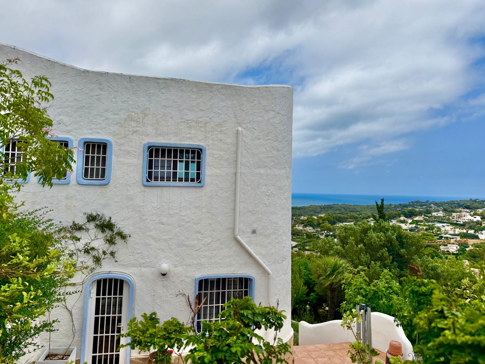 Fachada de estilo ibicenco con muros blancos texturizados, marcos azules y ventanas con rejas. La posición elevada ofrece vistas panorámicas al mar y al entorno natural de Jávea.