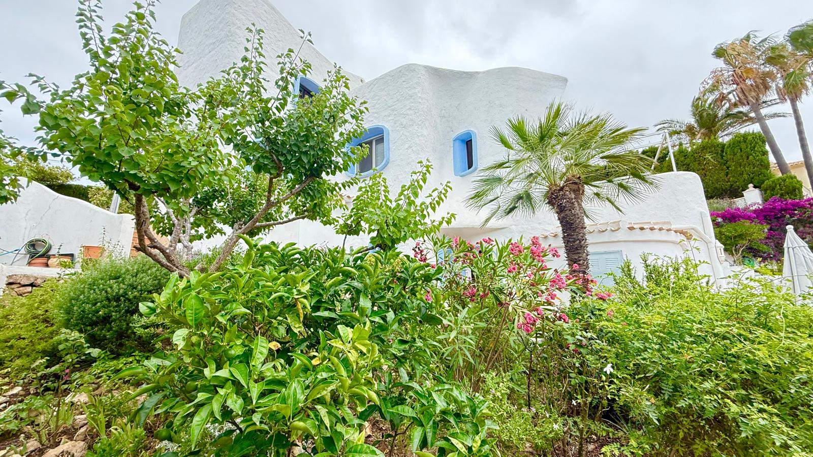 Fachada de estilo ibicenco con muros blancos texturizados y carpintería azul, rodeada de un jardín mediterráneo maduro con palmeras y arbustos en flor.