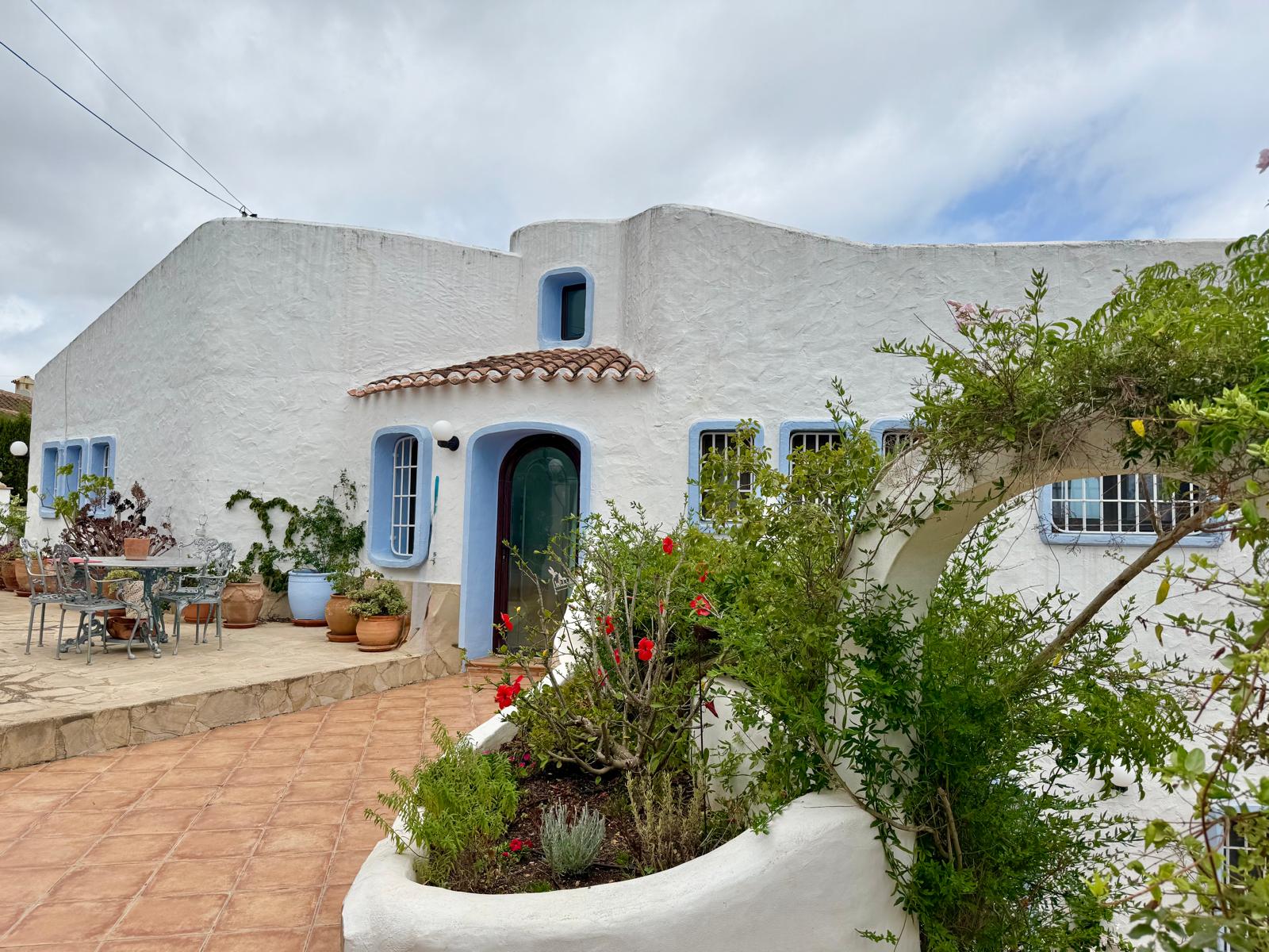 Fachada de estilo ibicenco con muros blancos y carpintería azul, destacando una terraza de baldosas de barro y jardineras integradas con vegetación mediterránea.