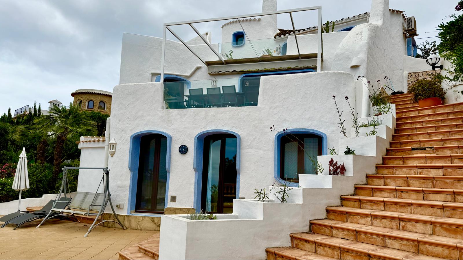 Fachada de estilo ibicenco con muros blancos y marcos azules. Los niveles están conectados por escaleras de barro cocido junto a terrazas con barandillas de cristal.