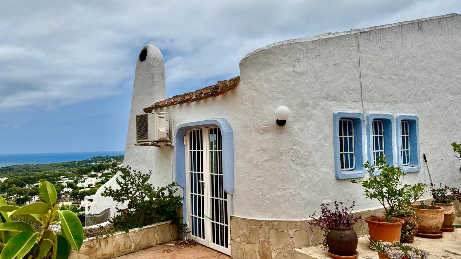 Fachada de estilo ibicenco con muros curvos blancos y marcos azules. Destaca la chimenea escultural y la terraza con macetas de barro frente a las vistas al mar.