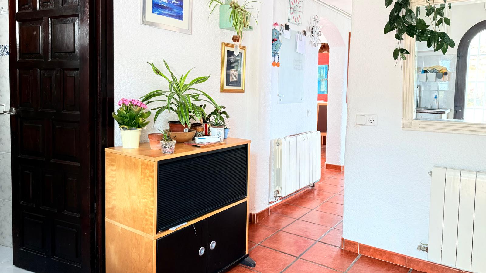 Pasillo de entrada con suelos de terracota, paredes blancas texturizadas y radiador de calefacción. Incluye una puerta de madera oscura y un arco que conecta con la zona de día.