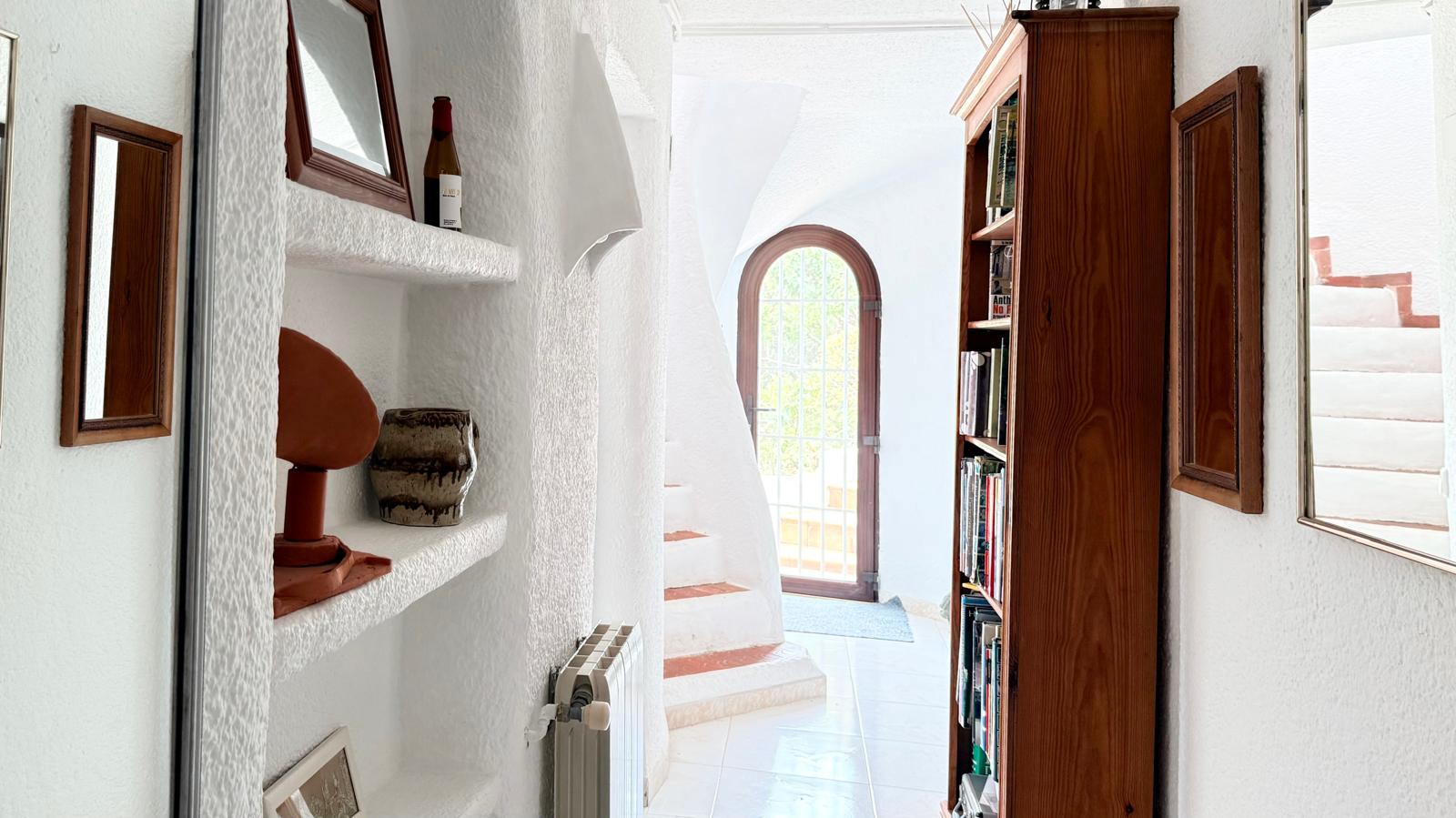 Pasillo de estilo ibicenco con paredes de yeso texturizado, estanterías integradas de obra y una escalera curva con peldaños de terracota que conduce a la planta superior.