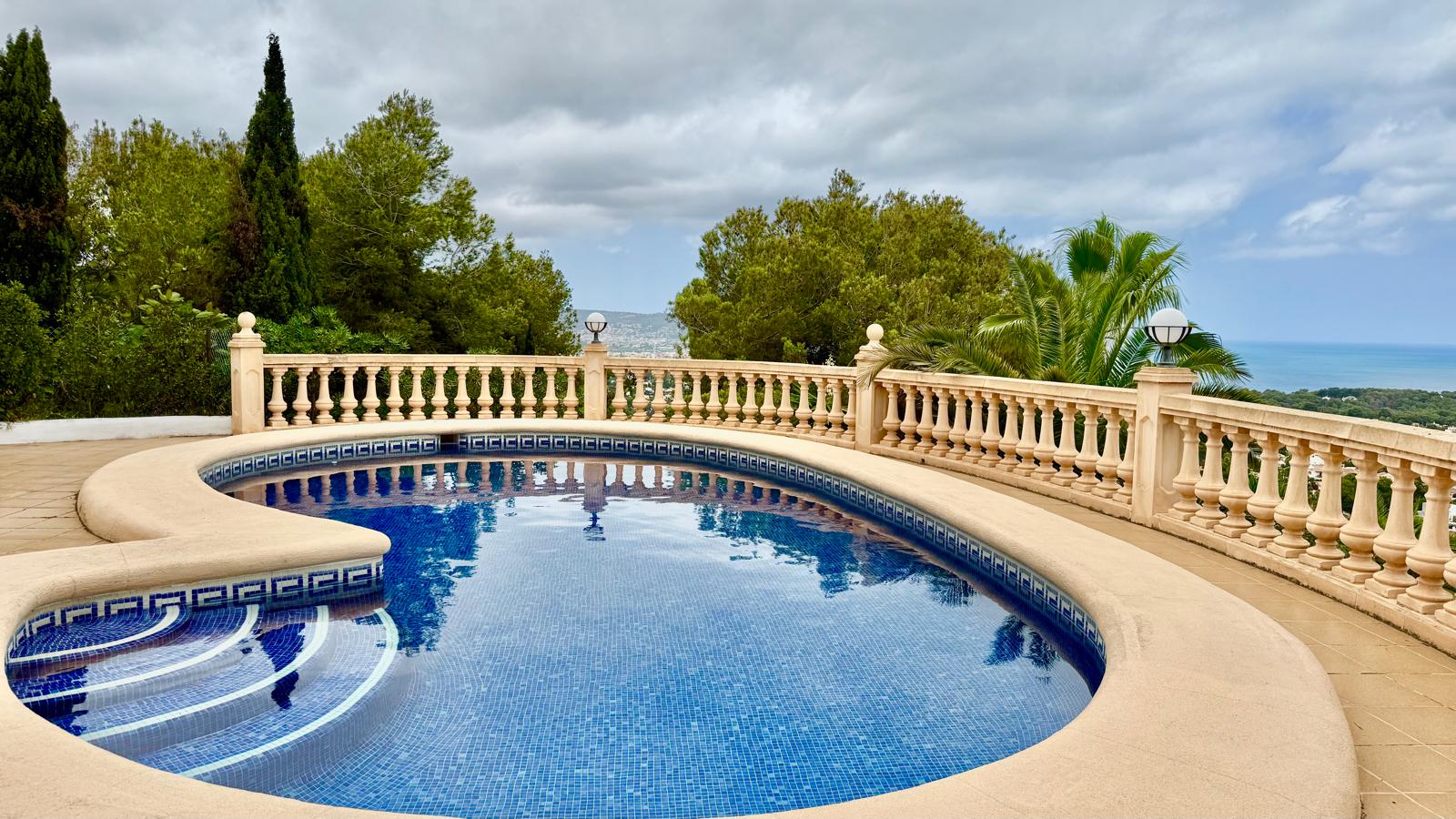 Piscina privada de forma curva con revestimiento de mosaico azul, rodeada por una terraza de piedra con balaustrada clásica y vistas panorámicas al mar y al entorno boscoso.