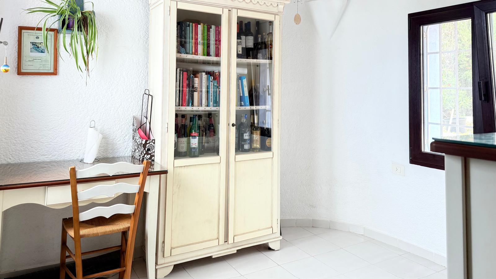 Rincón de despacho con escritorio clásico, silla de madera y vitrina blanca de estilo tradicional sobre suelos cerámicos claros y paredes blancas texturizadas.