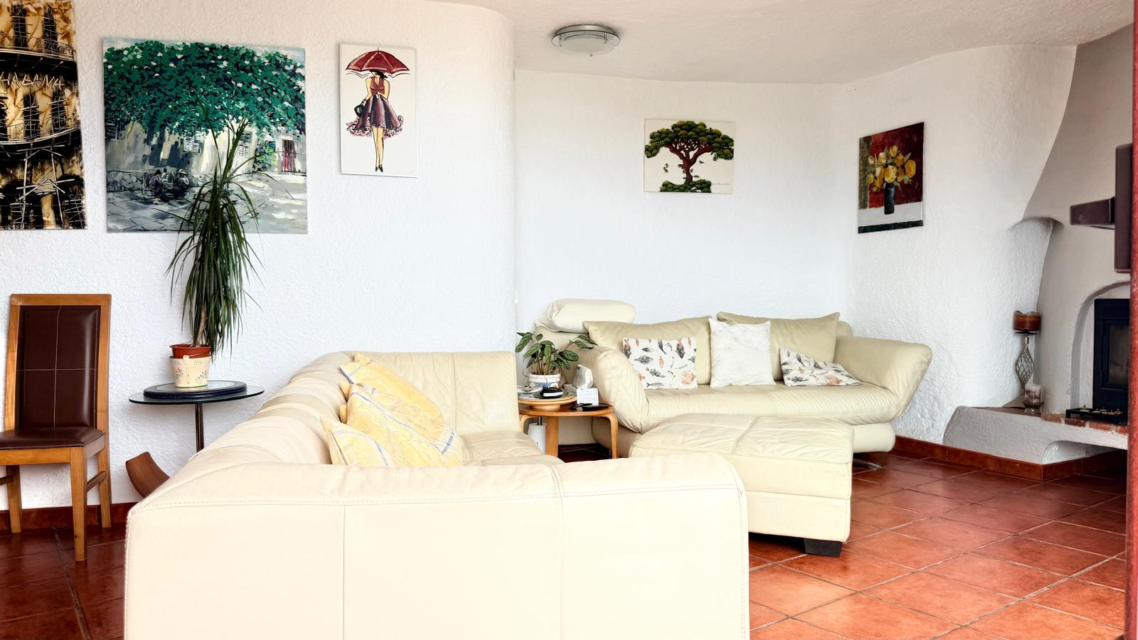 Salón acogedor con chimenea de leña, paredes blancas de estilo ibicenco, suelos de terracota y sofás de cuero en tonos claros bajo una arquitectura de líneas curvas.