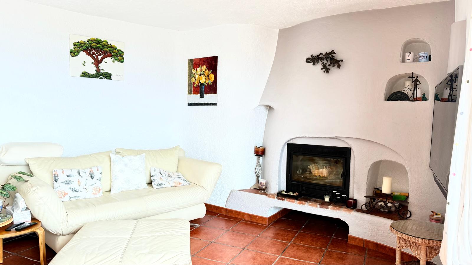 Salón de estilo ibicenco con chimenea de leña integrada, paredes blancas curvas y hornacinas decorativas. El suelo de baldosas de terracota aporta calidez al espacio principal.