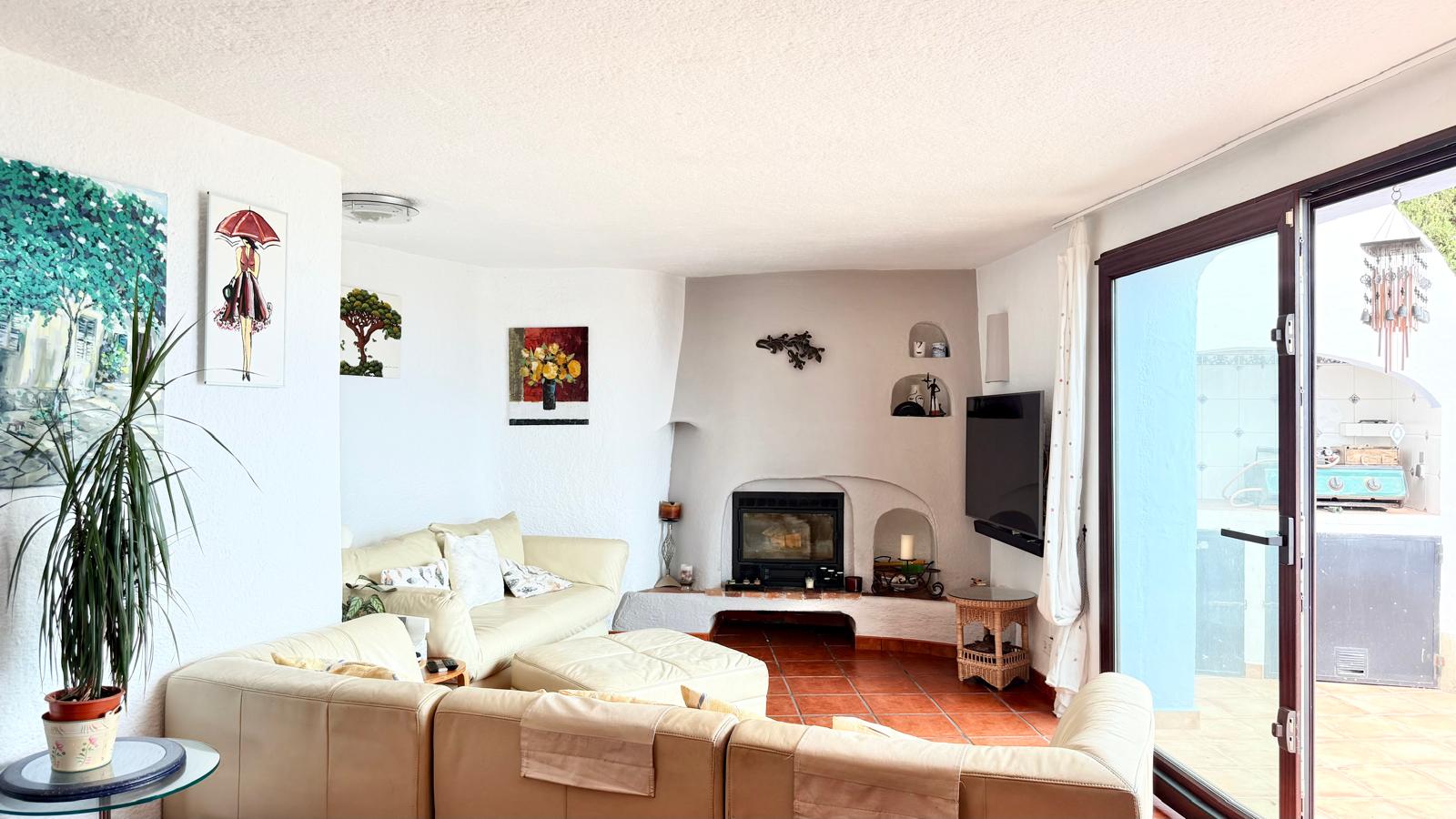Salón de estilo ibicenco con chimenea de leña, suelos de terracota y paredes curvas. Un gran ventanal conecta el espacio interior con la terraza exterior.
