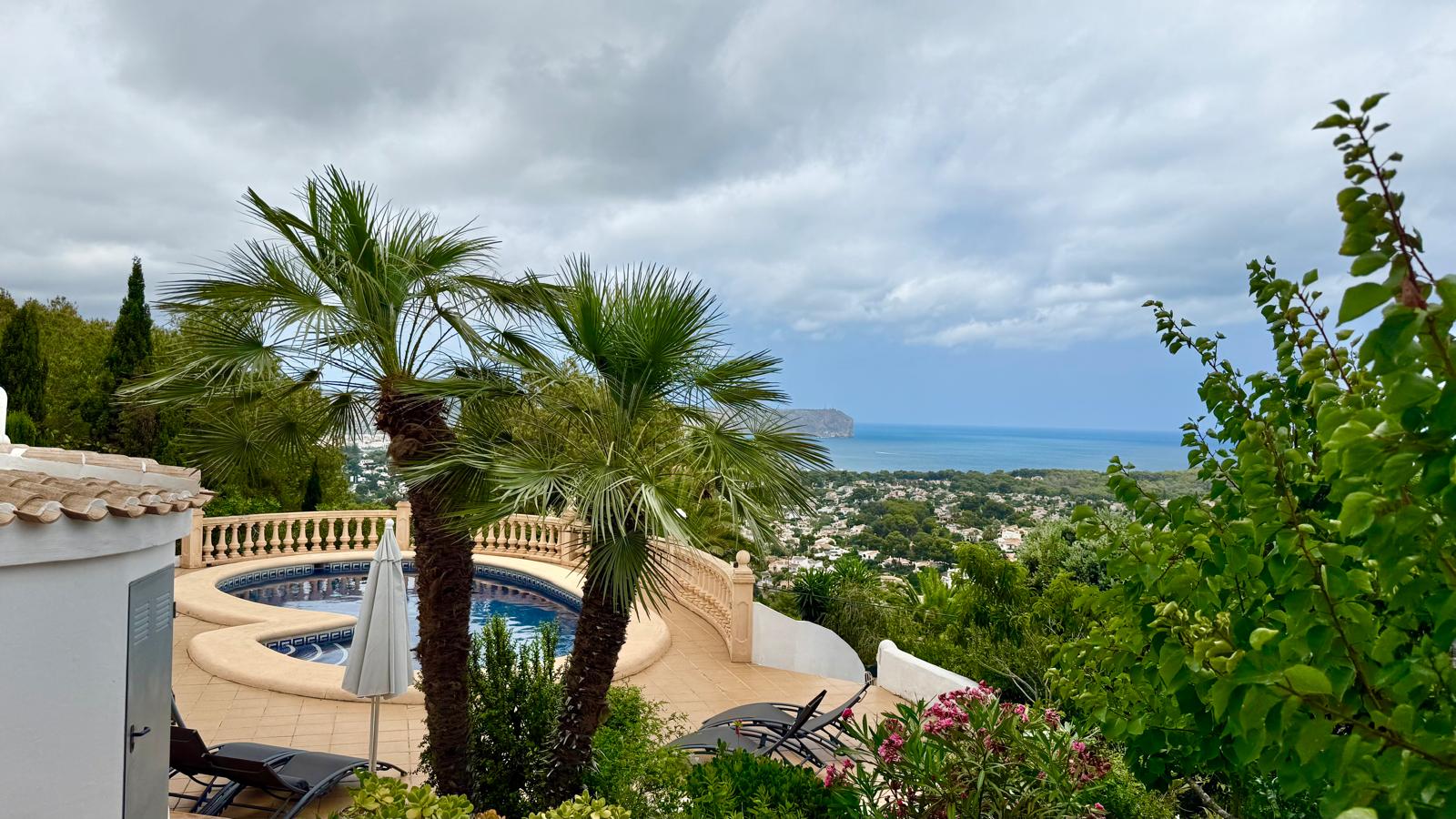 Vista elevada de la piscina privada rodeada de palmeras y vegetación mediterránea, con vistas panorámicas al mar y a la bahía de Jávea.