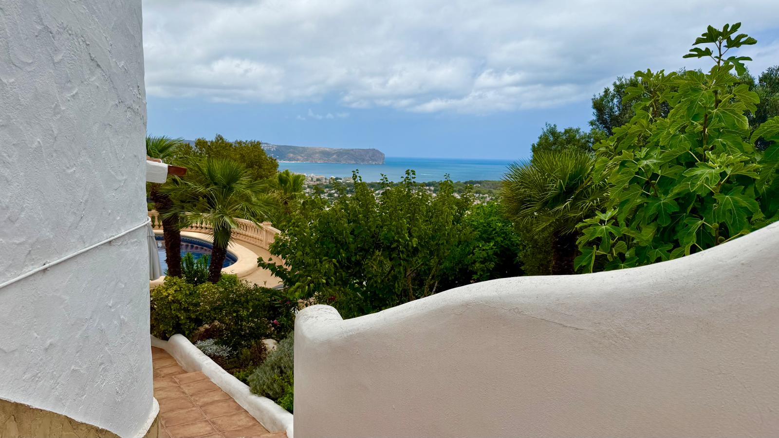 Vistas panorámicas al mar y la costa de Jávea desde las escaleras exteriores, flanqueadas por muros blancos curvos y vegetación mediterránea madura.