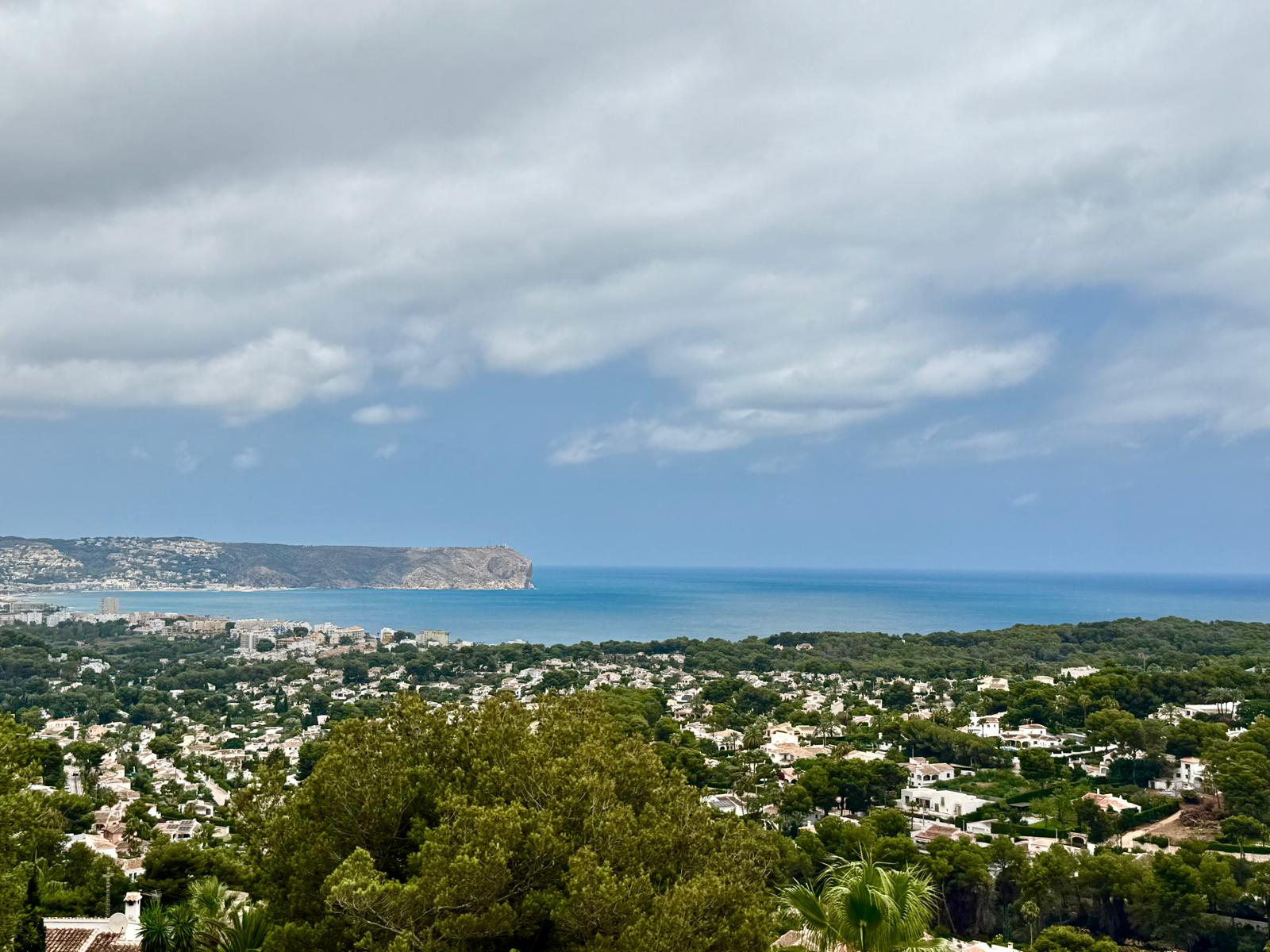 Vistas panorámicas elevadas hacia la bahía de Jávea y el mar Mediterráneo, destacando el perfil del cabo y el entorno residencial rodeado de vegetación autóctona.