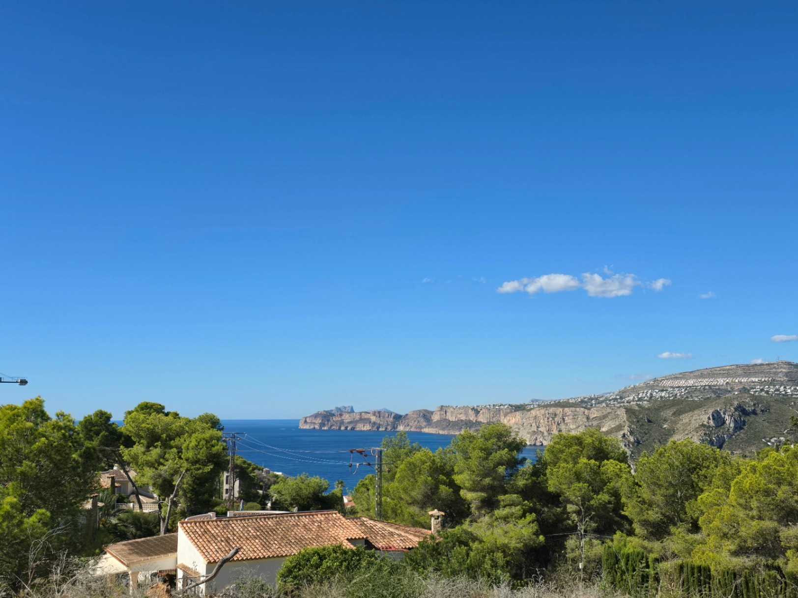 Vistas panorámicas al mar Mediterráneo y a los acantilados de Jávea, rodeadas de pinos autóctonos y tejados tradicionales bajo un cielo despejado.