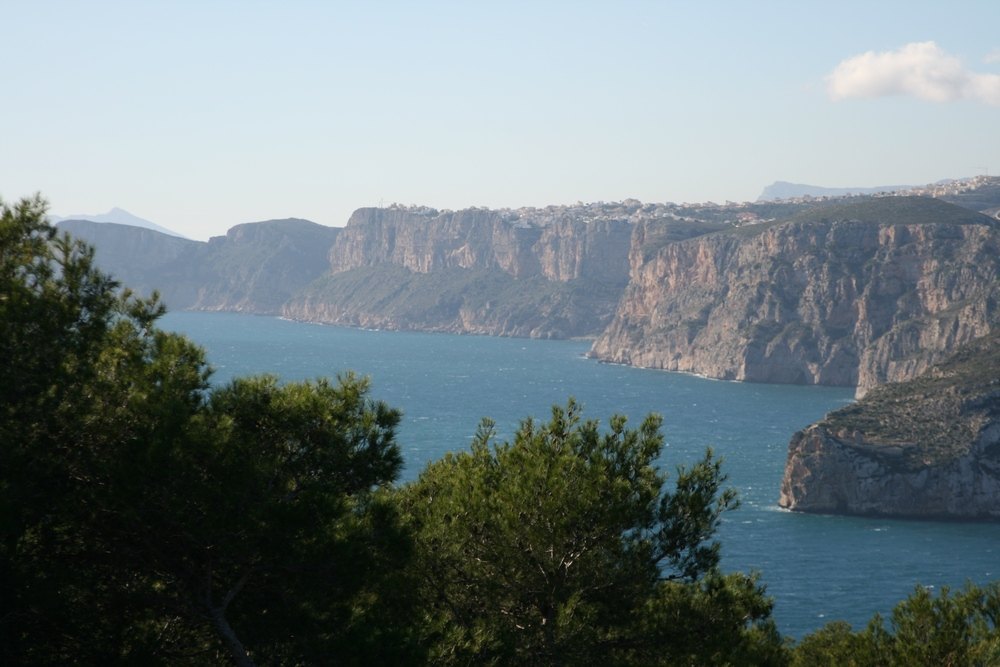 Vistas panorámicas al mar Mediterráneo y a los acantilados de la costa de Jávea, enmarcadas por vegetación autóctona de pinos bajo un cielo despejado.