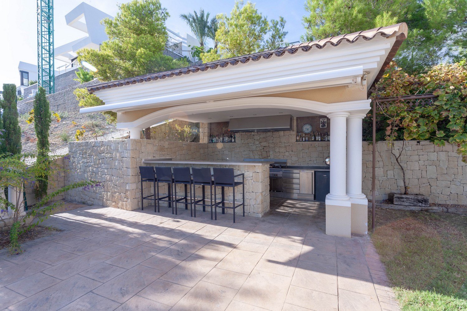Barbacoa exterior con barra y taburetes en villa de Benissa. Revestimiento de piedra y encimera de acero inoxidable.