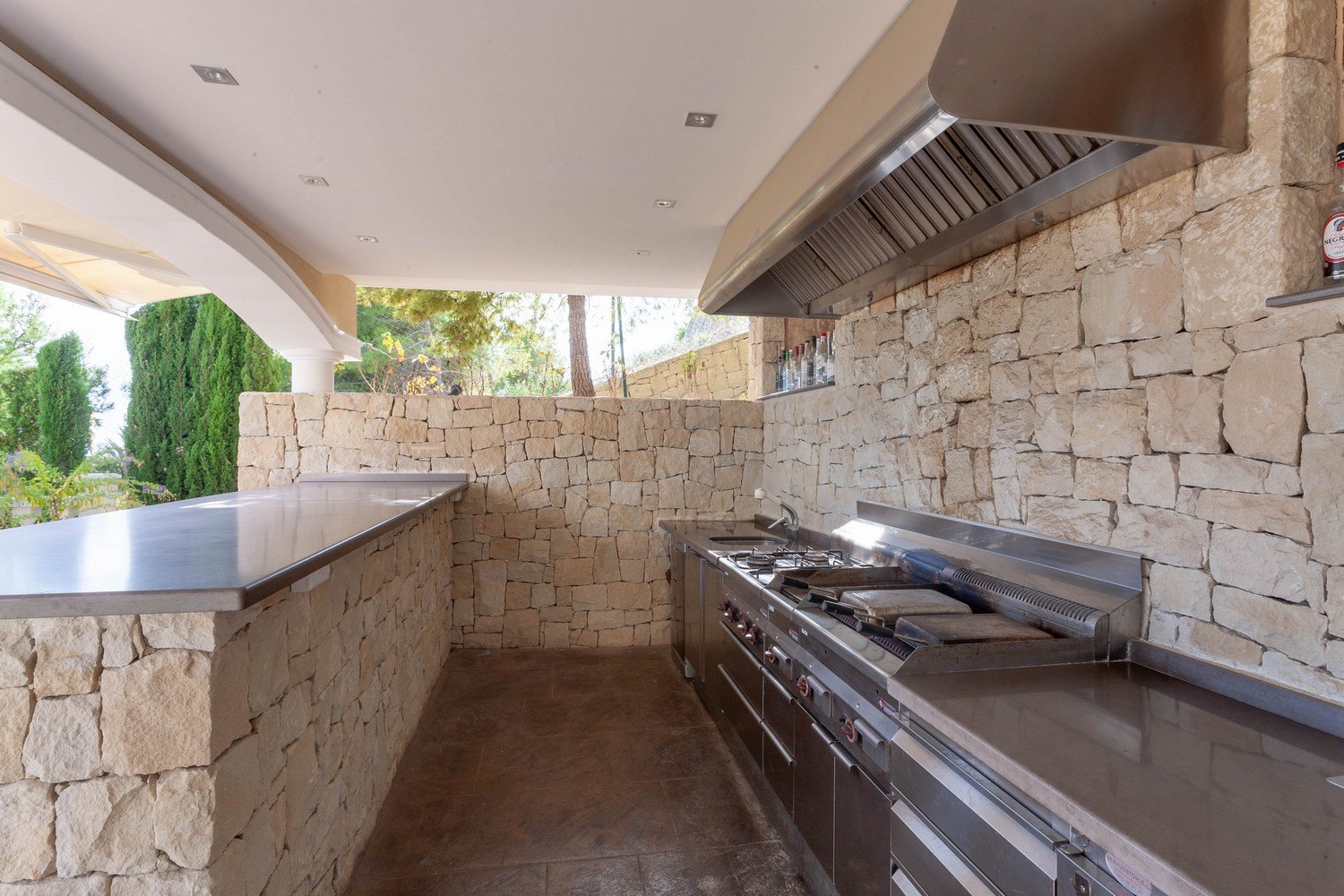 Cocina exterior con paredes de piedra, equipada con electrodomésticos de acero inoxidable y barra con encimera gris.