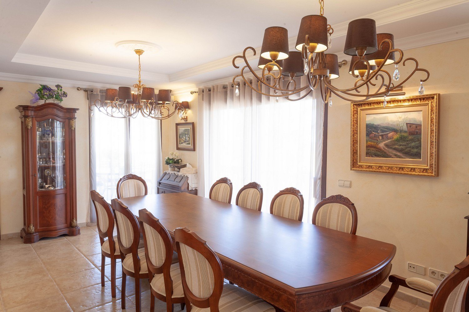 Comedor clásico con mesa de madera, sillas tapizadas y vitrina antigua. Iluminación cálida con lámparas de araña.