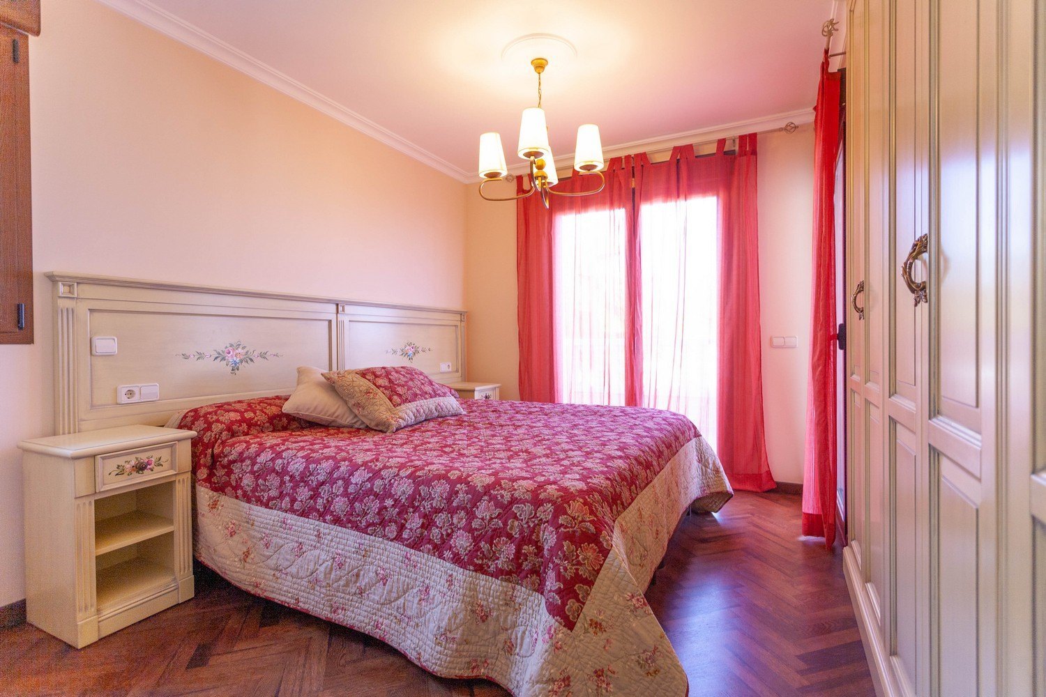 Dormitorio con cama de matrimonio, mesita de noche y armario de madera clara con detalles florales.