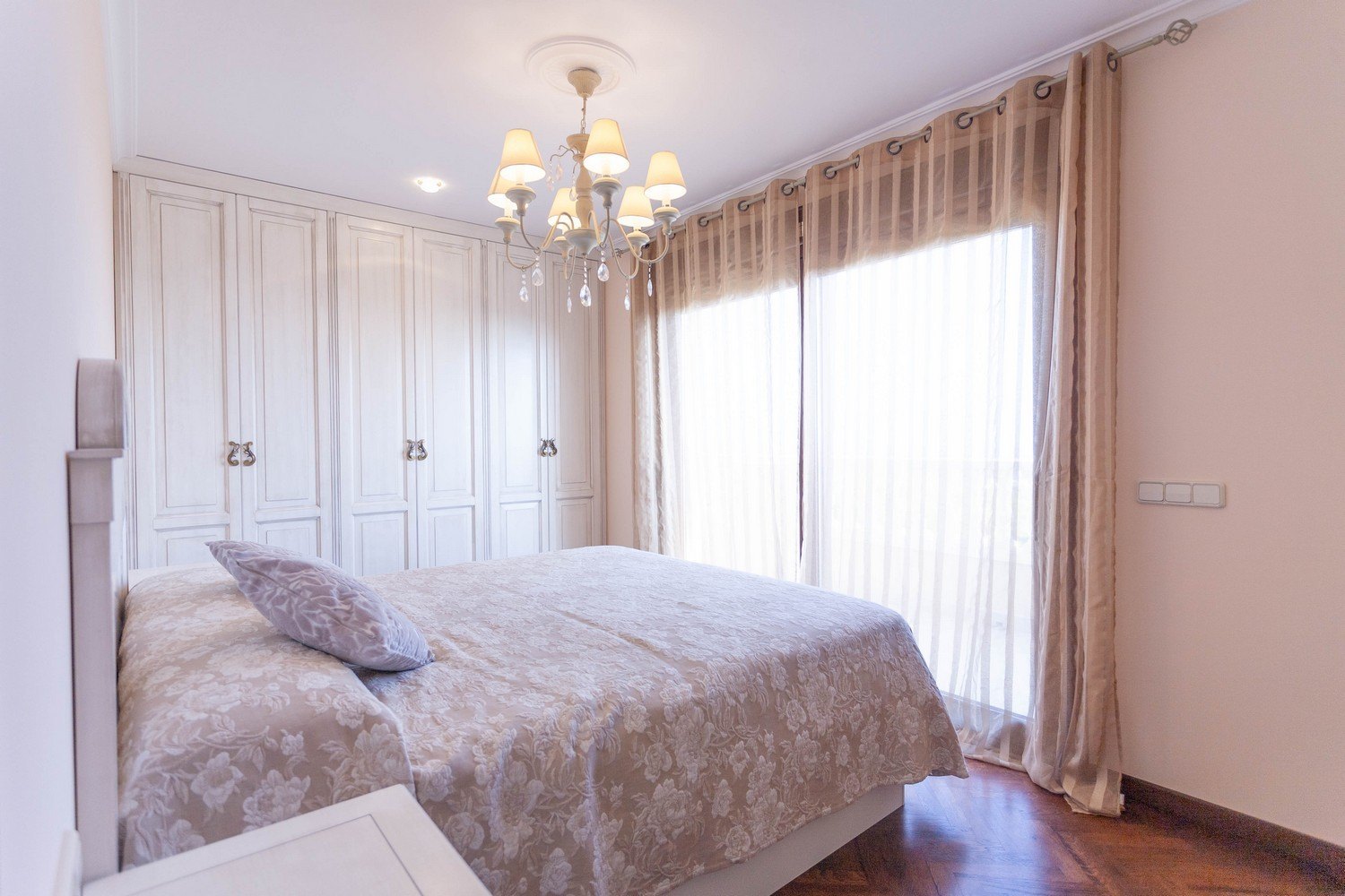 Dormitorio con cama doble, armario empotrado blanco, lámpara de araña y cortinas beige.
