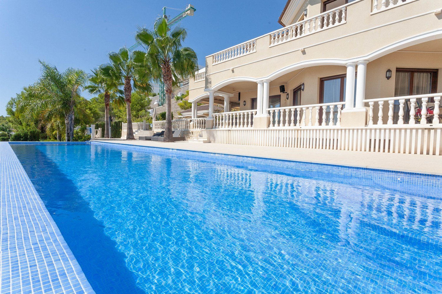 Vista de la piscina azul turquesa con borde de mosaico y la fachada de la villa de estilo mediterráneo.