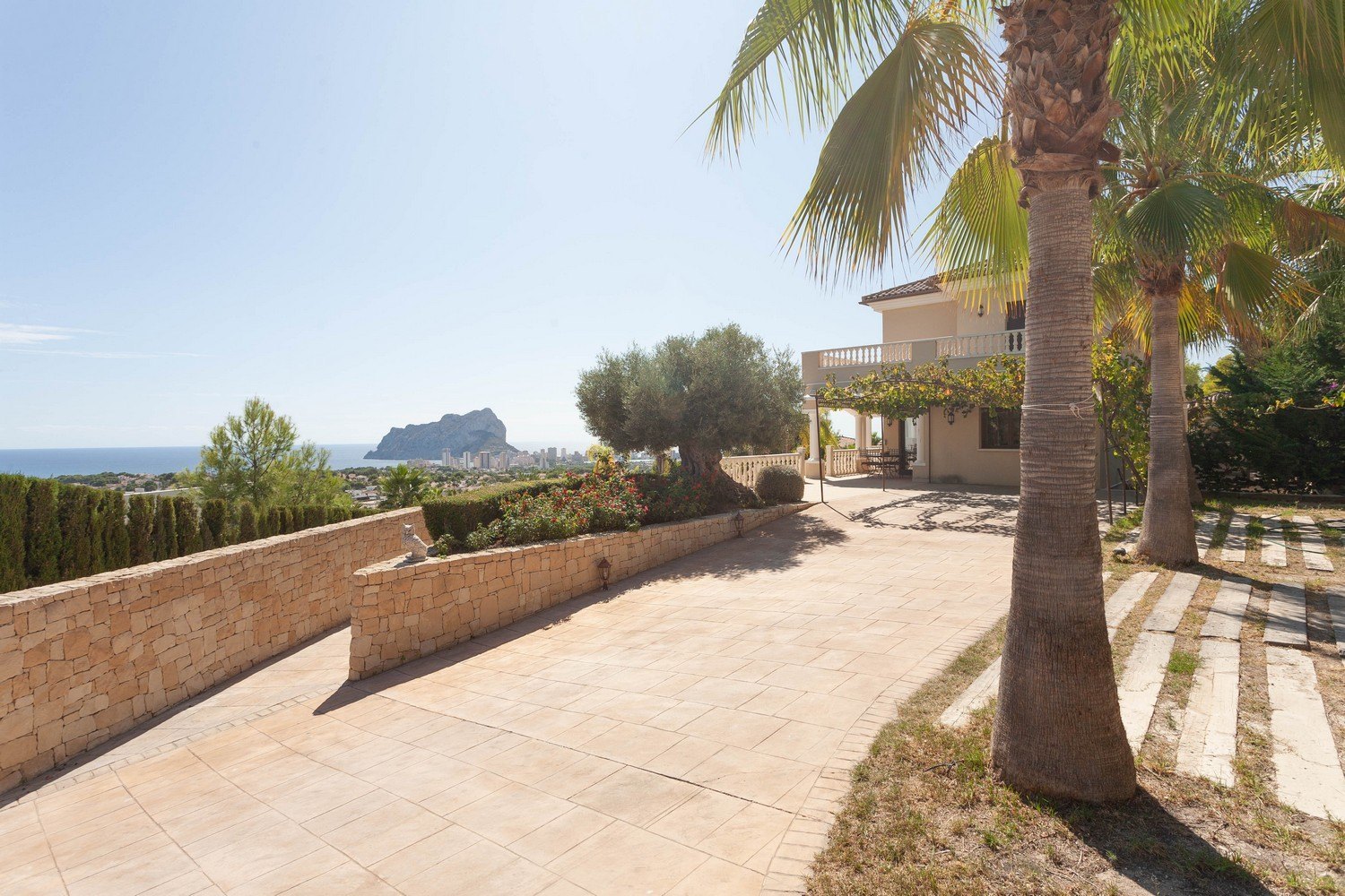 Vista exterior de la villa con camino de losas, muro de piedra y vistas al mar.