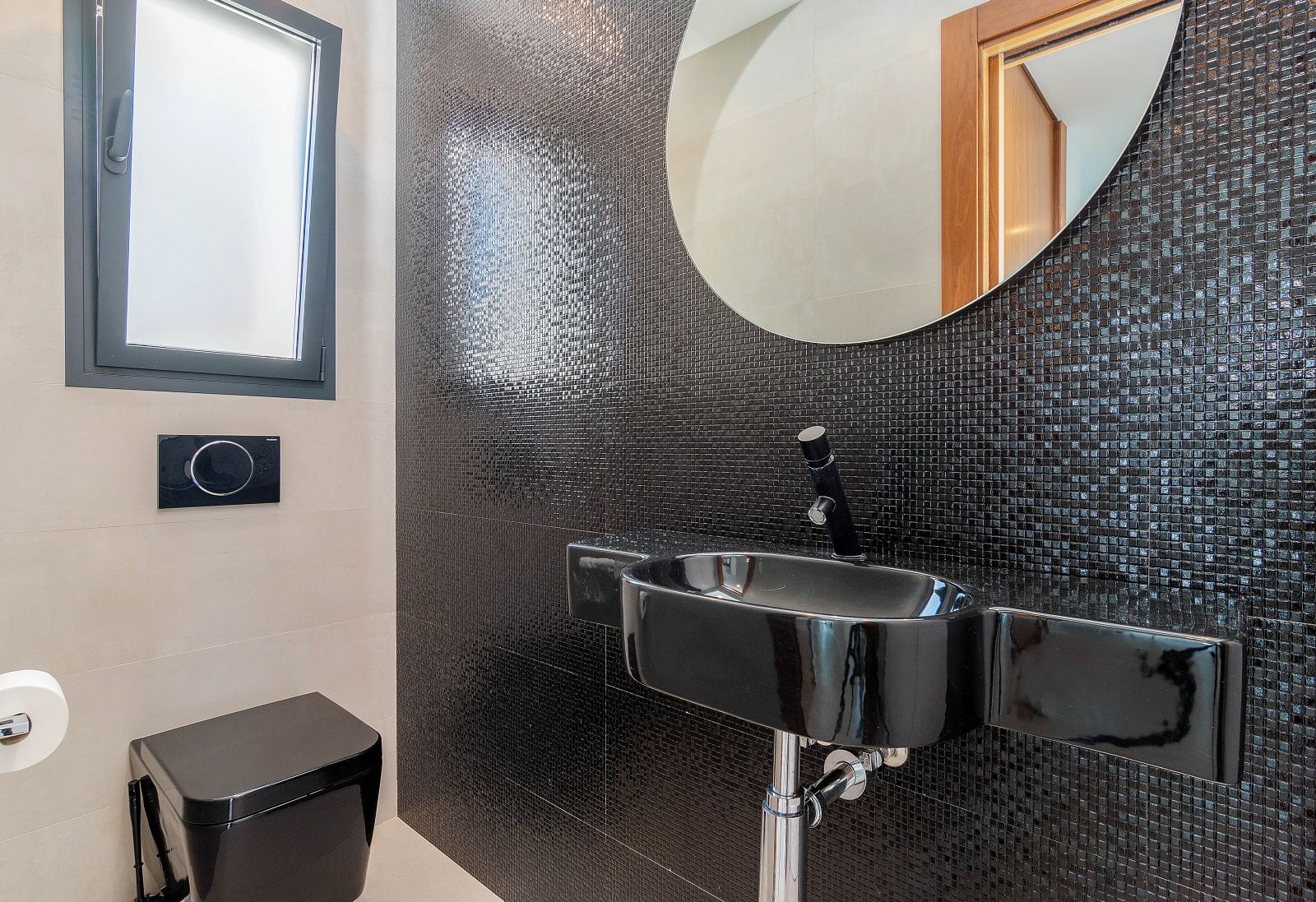 Aseo de cortesía con pared de mosaico negro, lavabo y sanitario suspendido en color negro, espejo circular y ventana de ventilación.