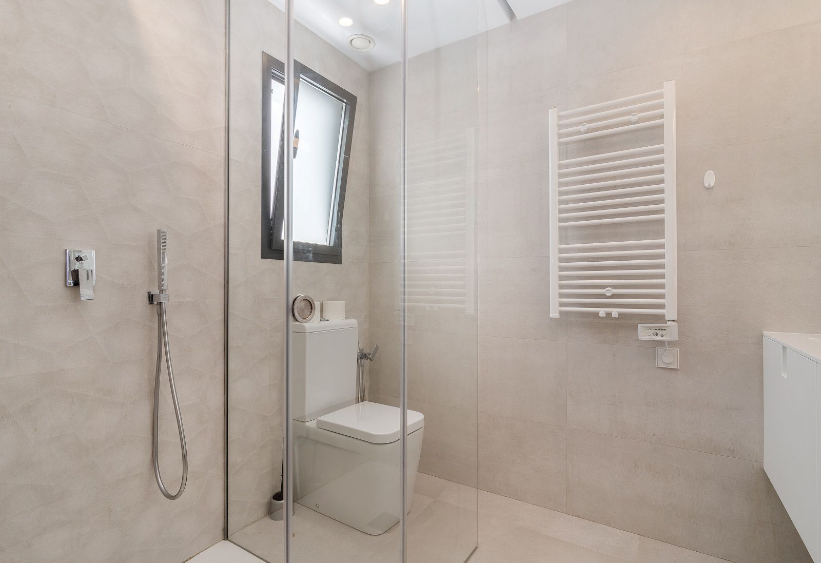 Baño moderno con ducha de obra, mampara de cristal, inodoro blanco y radiador toallero eléctrico sobre paredes de gres con relieve en tonos arena.