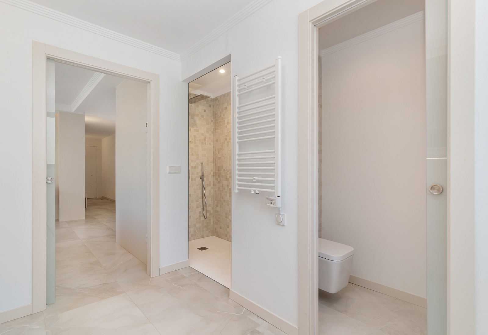Baño moderno con ducha de obra revestida en mosaico beige, radiador toallero blanco y sanitario suspendido. Suelos de gres porcelánico y puertas correderas de cristal.