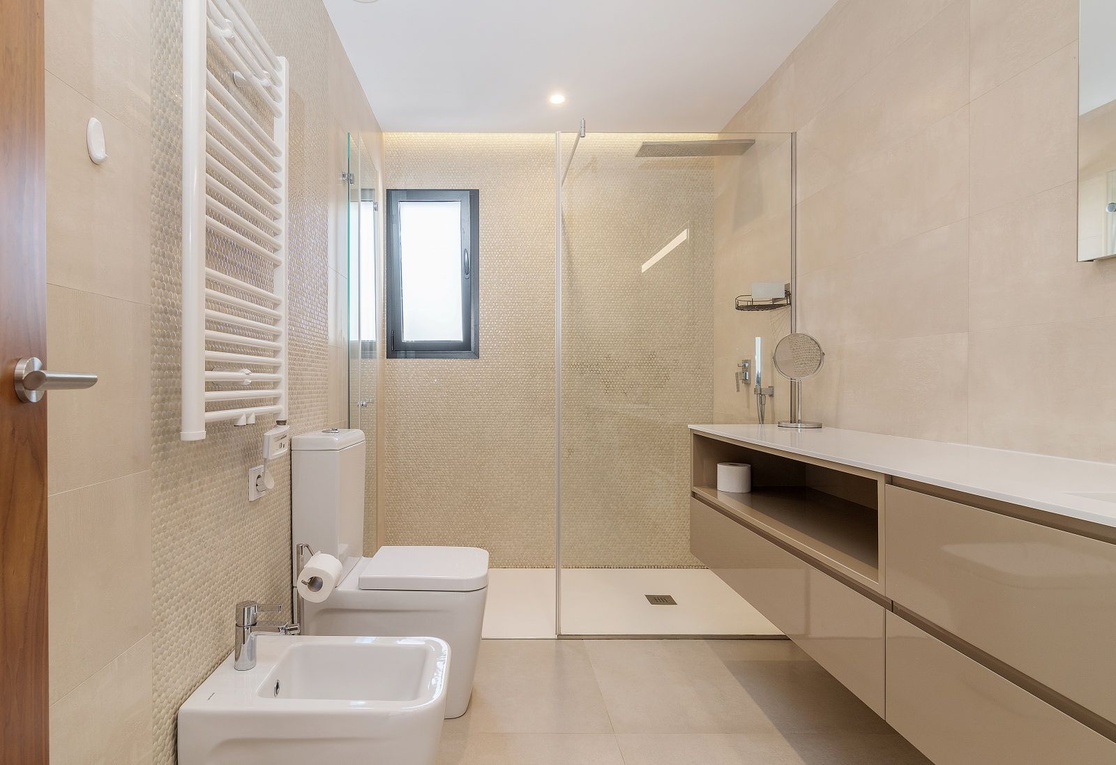 Baño moderno con ducha italiana, mampara de cristal, mueble suspendido en tonos arena, bidé y radiador toallero blanco sobre paredes de gres cerámico.